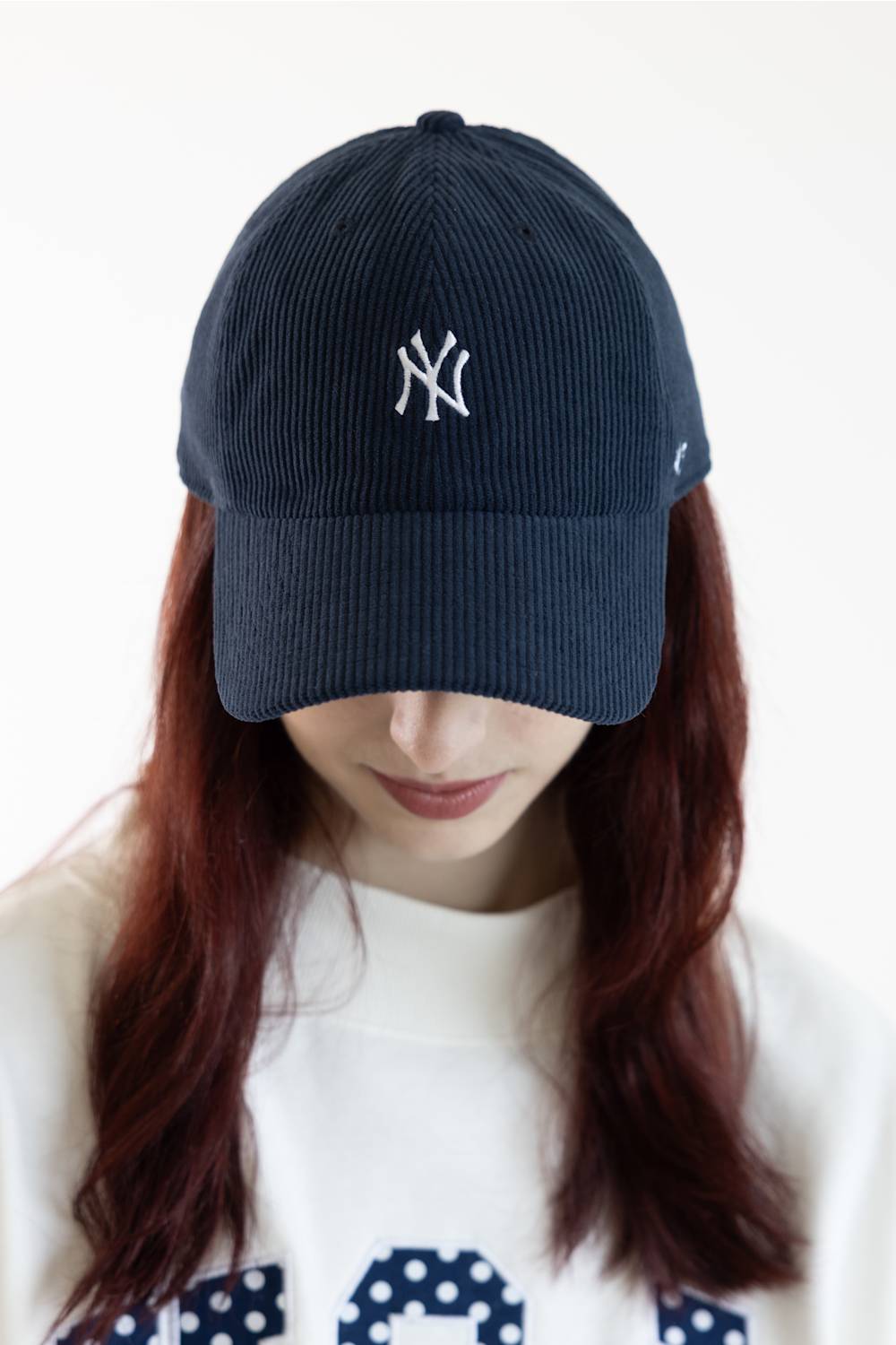 A person wearing a dark navy blue corduroy baseball cap with a white embroidered New York Yankees logo on the front, paired with a white sweatshirt. The background is a clean, bright white studio setting.