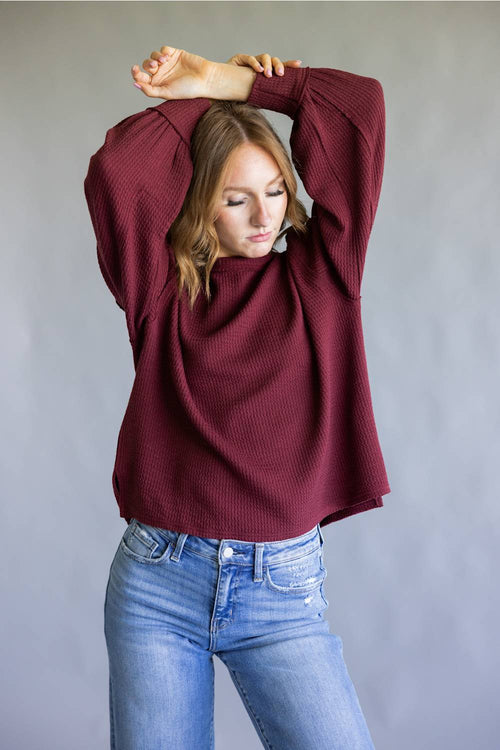 A woman wearing a maroon ribbed knit long-sleeve sweater and blue jeans, posing with her arms raised against a plain gray studio background.