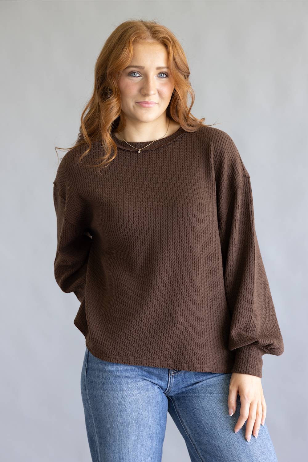 A woman with red hair wearing a brown waffle-knit long-sleeve sweater and blue jeans, standing against a neutral gray studio background.