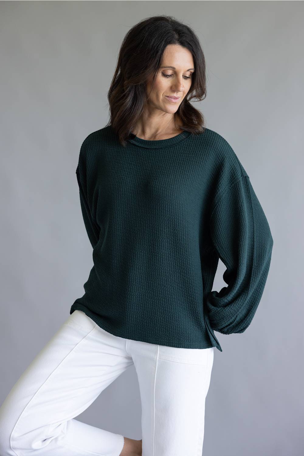 A woman wearing a dark forest green ribbed knit sweater with balloon sleeves and white pants, posing against a neutral gray studio background.