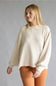 A woman wearing a cream-colored waffle-knit long-sleeve sweater and rust-orange pants, standing against a neutral gray studio background.