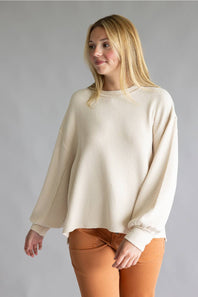 A woman wearing a cream-colored waffle-knit long-sleeve sweater and rust-orange pants, standing against a neutral gray studio background.