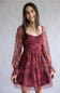 A woman wearing a burgundy and pink floral tiered dress with sheer long sleeves, standing against a neutral gray studio background.