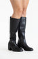 A pair of black leather knee-high boots with block heels, shown on a model's feet against a plain white studio background.