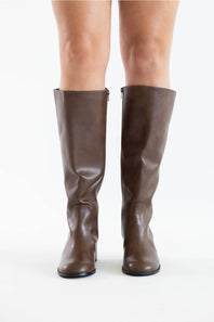 A pair of knee-high brown faux-leather boots with a low block heel, shown on a model's feet against a plain white background.