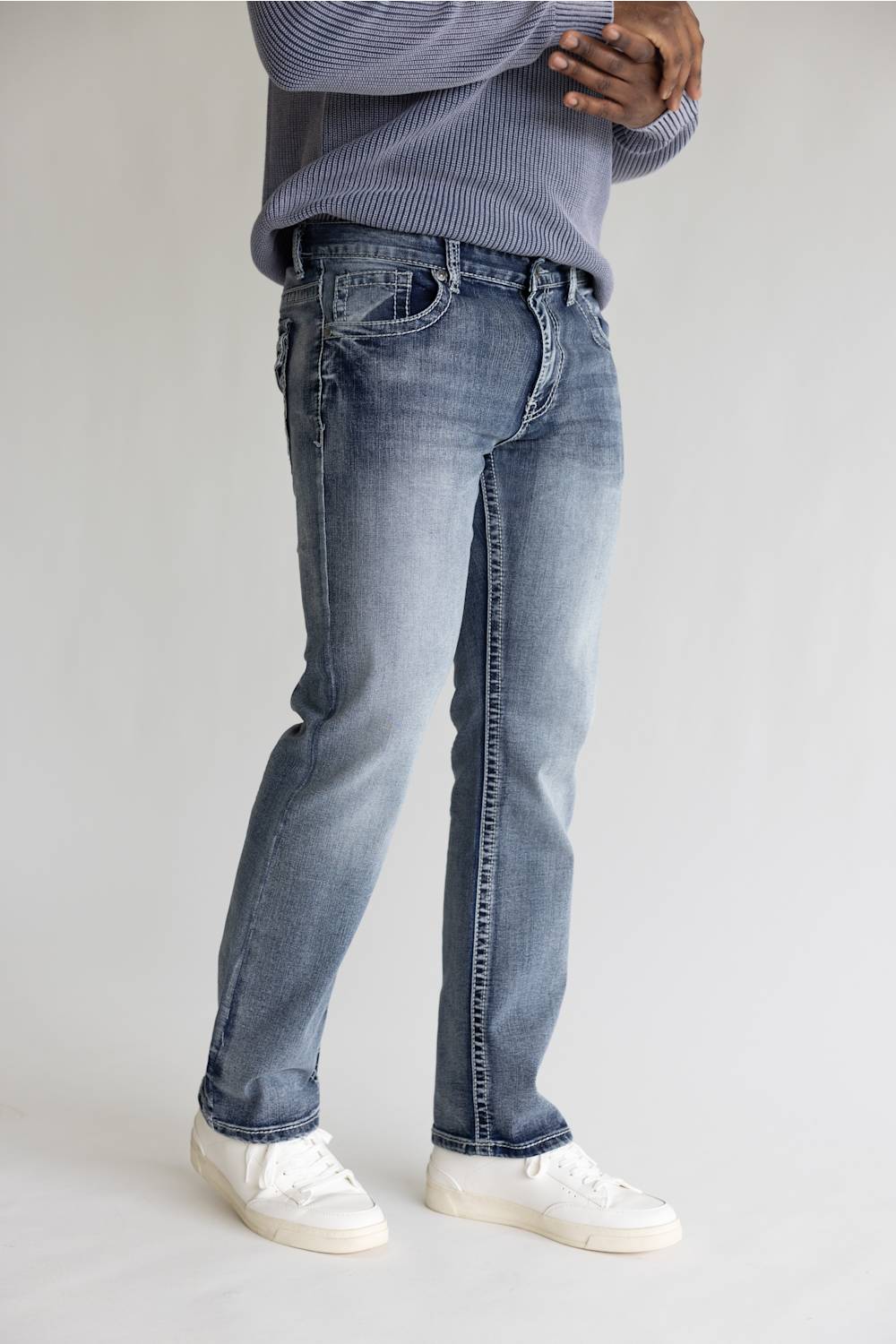 A person wearing light-wash blue denim jeans with a faded wash, paired with a gray ribbed sweater and white sneakers, standing against a light gray studio background.