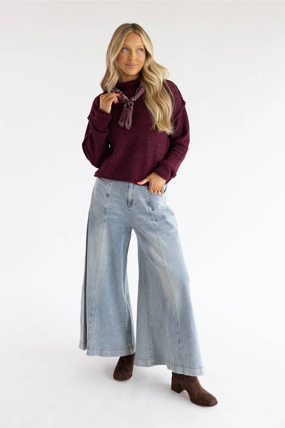 A woman modeling a dark burgundy knit sweater, wide-leg light-wash blue jeans, and brown ankle boots against a plain white studio background.