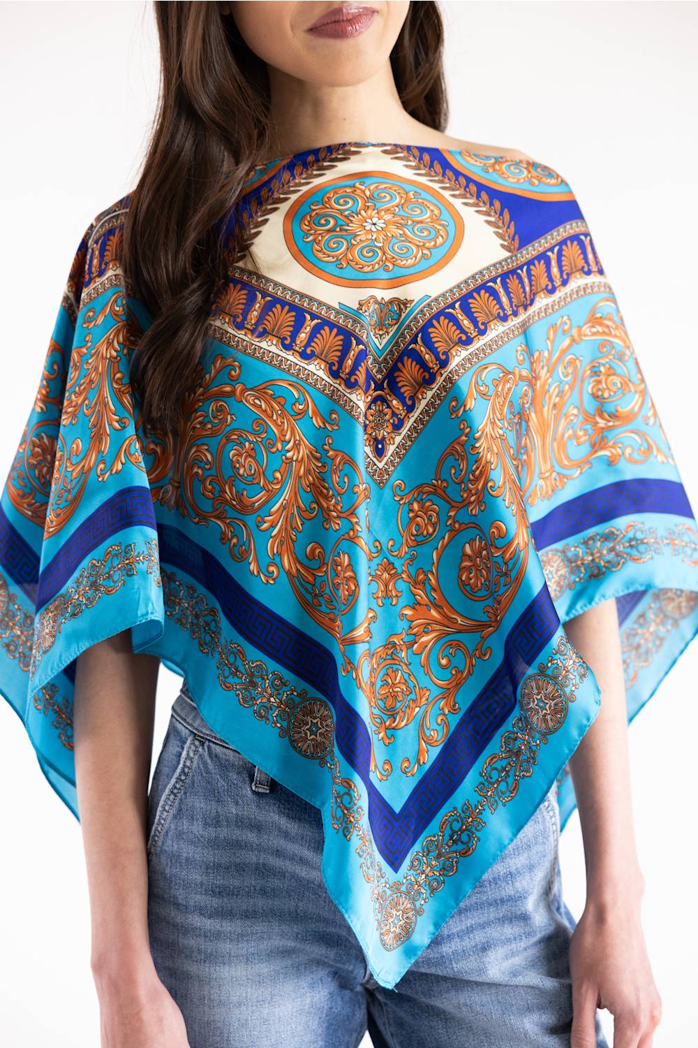 A woman wearing a bright turquoise and blue patterned poncho-style top with ornate gold and brown paisley motifs, paired with blue jeans, against a plain white background.