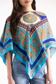 A woman wearing a bright turquoise and blue patterned poncho-style top with ornate gold and brown paisley motifs, paired with blue jeans, against a plain white background.