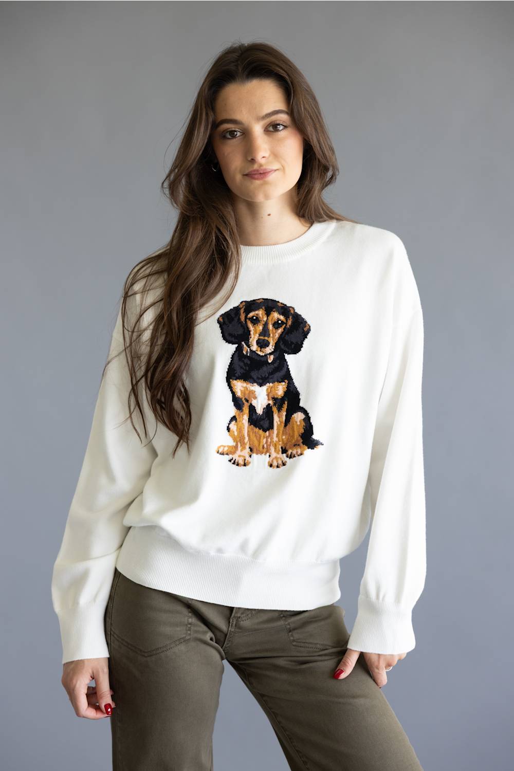 A woman modeling a white crewneck sweatshirt featuring a detailed graphic of a black and tan dachshund on the front, paired with olive green pants against a solid gray studio background.