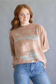 A woman with red hair wearing a faded brown graphic t-shirt with 'BIBBABONI' and 'MAGIC SUNSET UNDERWAGE' printed in light blue, paired with blue jeans, against a solid gray background.