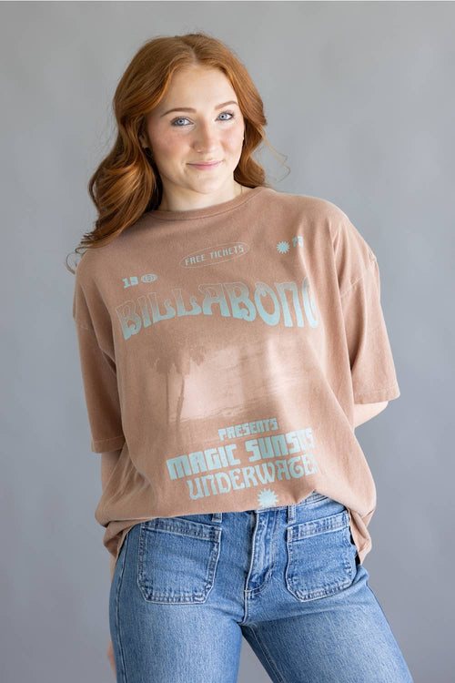 A woman with red hair wearing a faded brown graphic t-shirt with 'BIBBABONI' and 'MAGIC SUNSET UNDERWAGE' printed in light blue, paired with blue jeans, against a solid gray background.