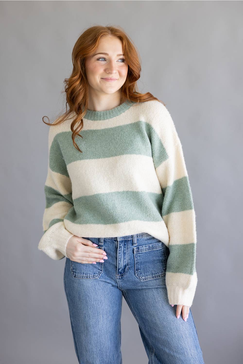 A woman with red hair wearing a fuzzy knit sweater with horizontal sage green and cream stripes, paired with blue jeans, standing against a solid gray studio background.