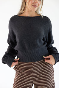 A woman wearing a dark charcoal gray knit sweater with a wide boat neckline and ribbed hem, paired with brown and beige striped pants, against a plain white background.