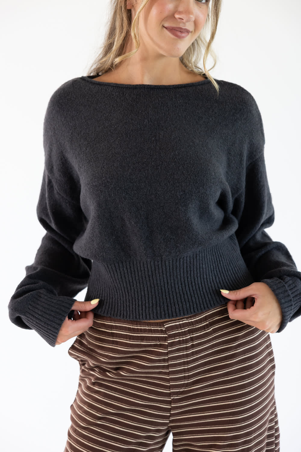 A woman wearing a dark charcoal gray knit sweater with a wide boat neckline and ribbed hem, paired with brown and beige striped pants, against a plain white background.