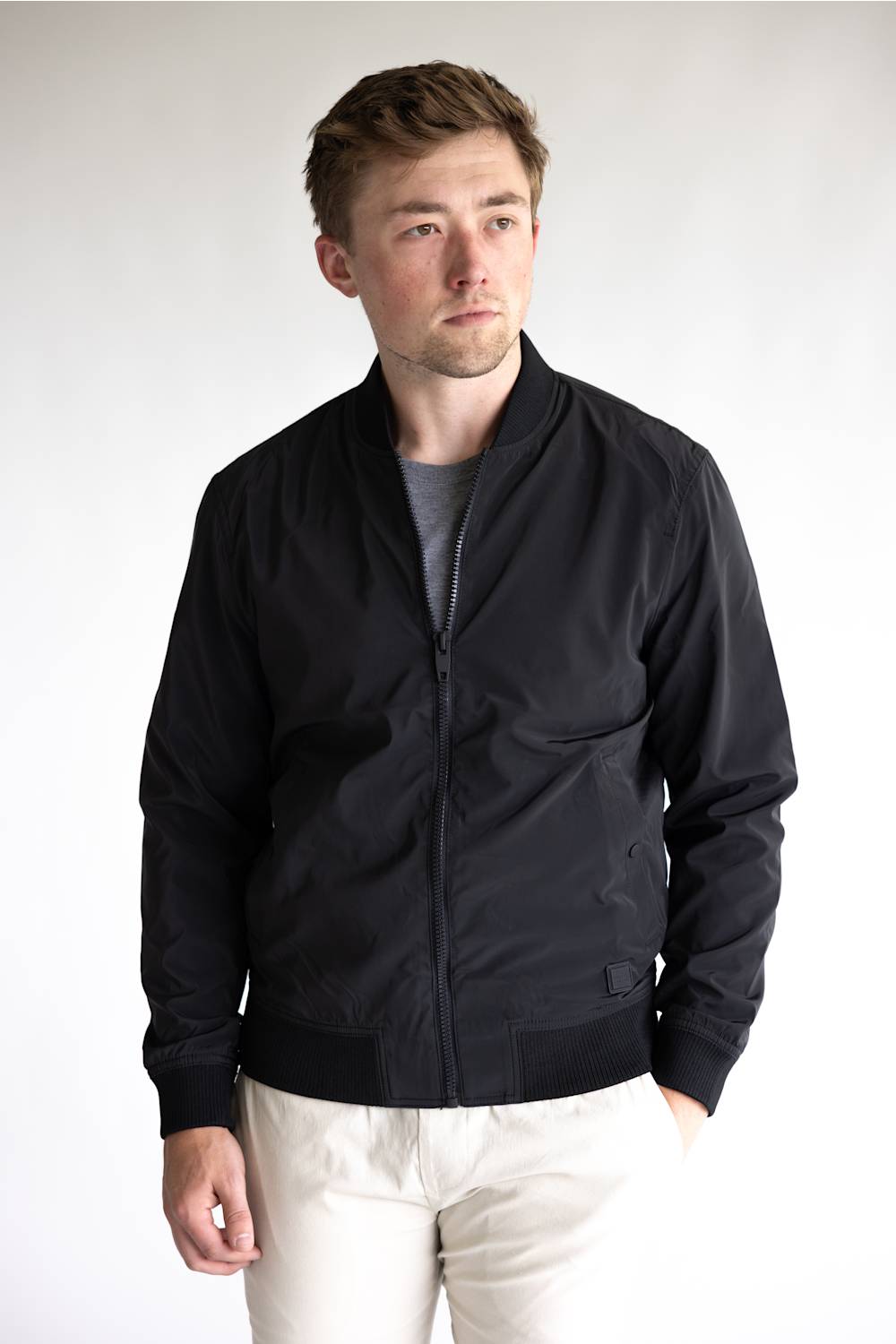 A man wearing a black zip-up bomber jacket over a gray t-shirt and off-white trousers, standing against a light gray studio background.
