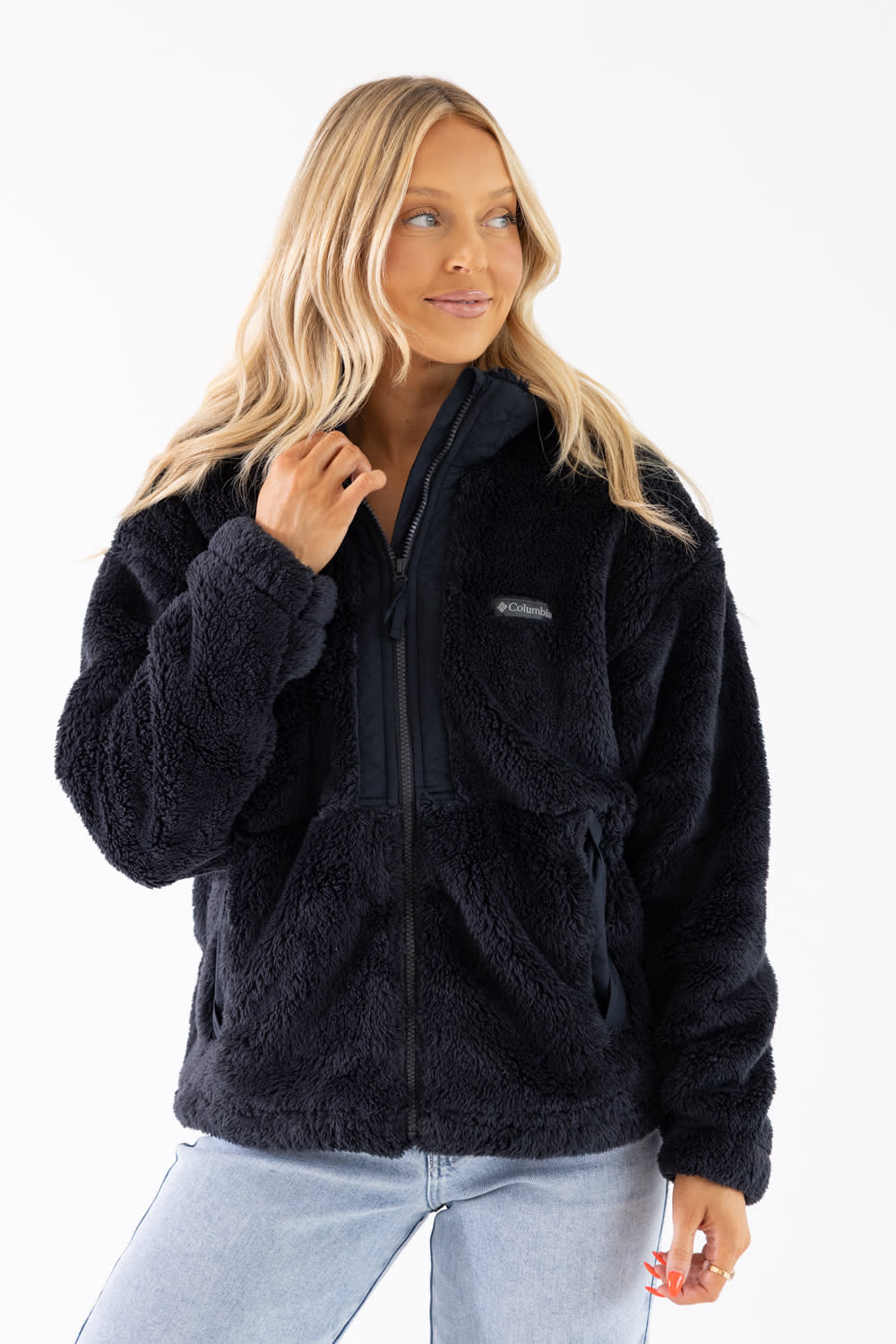 A woman wearing a black fleece zip-up jacket and light blue jeans, photographed against a plain white studio background.