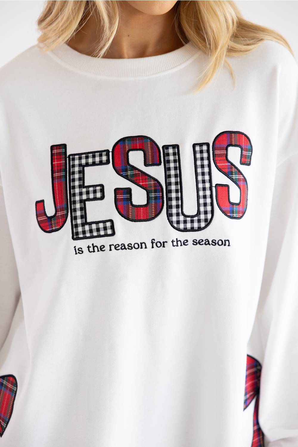 A close-up of a person wearing a white crewneck sweatshirt with a graphic that reads 'JESUS is the reason for the season' in black text, with the letters filled in red, black, and white plaid tartan patterns.