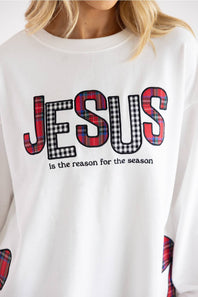 A close-up of a person wearing a white crewneck sweatshirt with a graphic that reads 'JESUS is the reason for the season' in black text, with the letters filled in red, black, and white plaid tartan patterns.