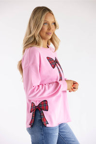 A woman modeling a pink crewneck sweatshirt with red and green plaid bow details on the chest and lower hem, paired with blue jeans against a plain white background.