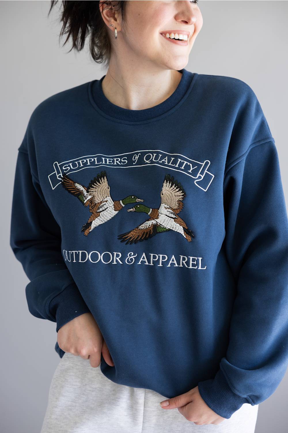 A woman wearing a navy blue crewneck sweatshirt with an embroidered graphic of two mallards and the text 'Suppliers of Quality Outdoors & Apparel'.