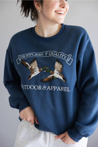 A woman wearing a navy blue crewneck sweatshirt with an embroidered graphic of two mallards and the text 'Suppliers of Quality Outdoors & Apparel'.