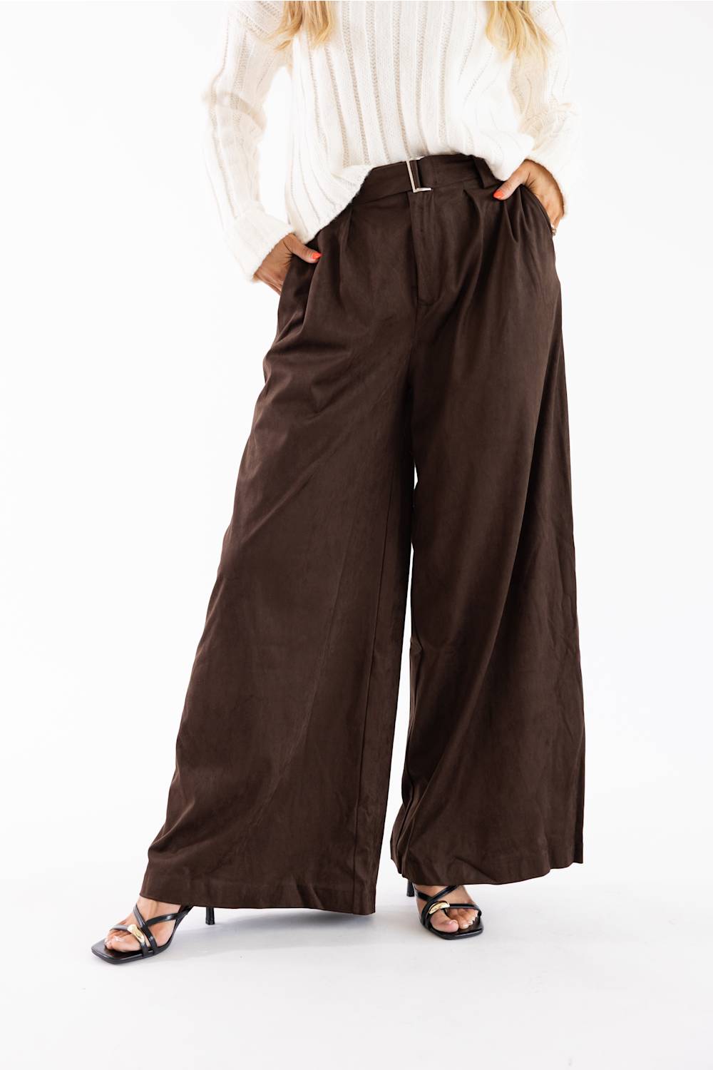 A model wearing high-waisted, wide-leg brown pants with a matching belt, paired with a white long-sleeve top and black heeled sandals, shown from the waist down on a white studio background.