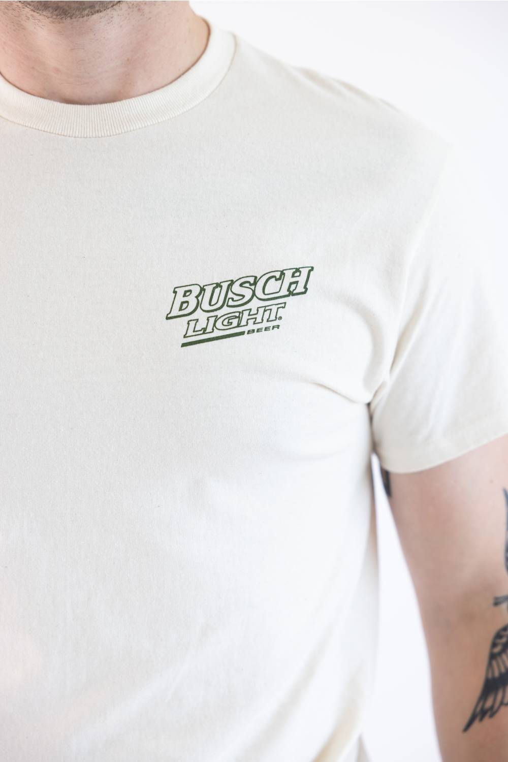 Close-up of a person wearing a cream-colored crewneck t-shirt with a green outlined 'BUSCH LIGHT' logo printed on the left chest.