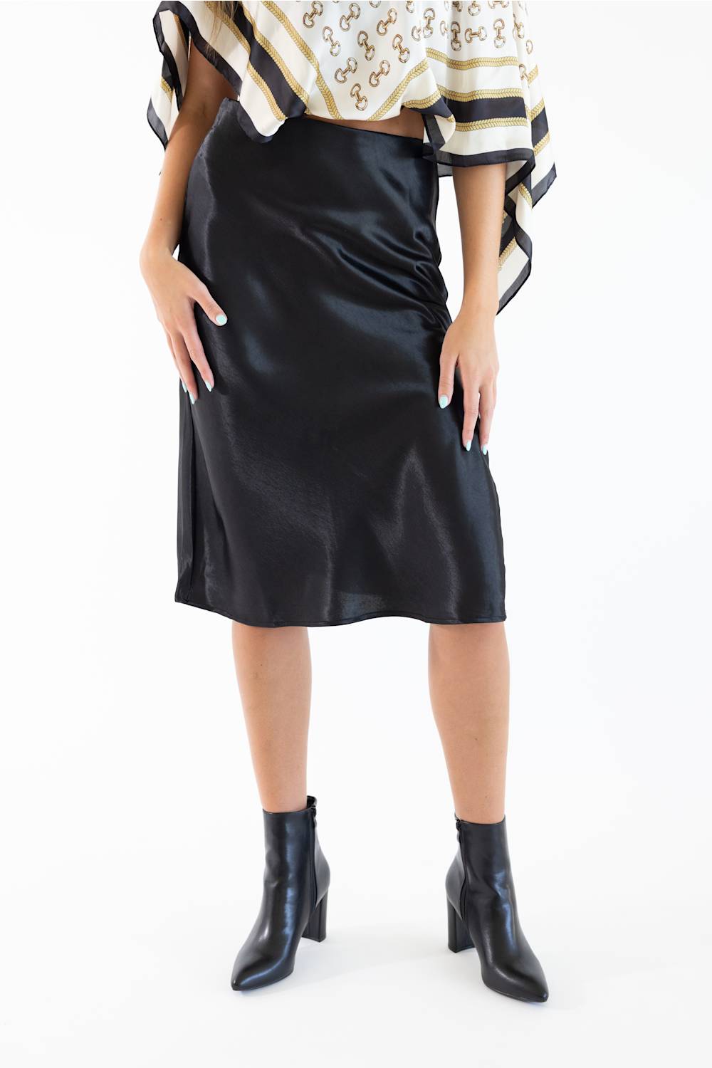 A model wearing a black satin midi skirt with a side slit, paired with a patterned top and black ankle boots, against a plain white background.
