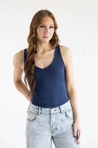 A woman with long wavy light brown hair wearing a navy blue tank top and light-wash blue jeans, standing against a plain white studio background.