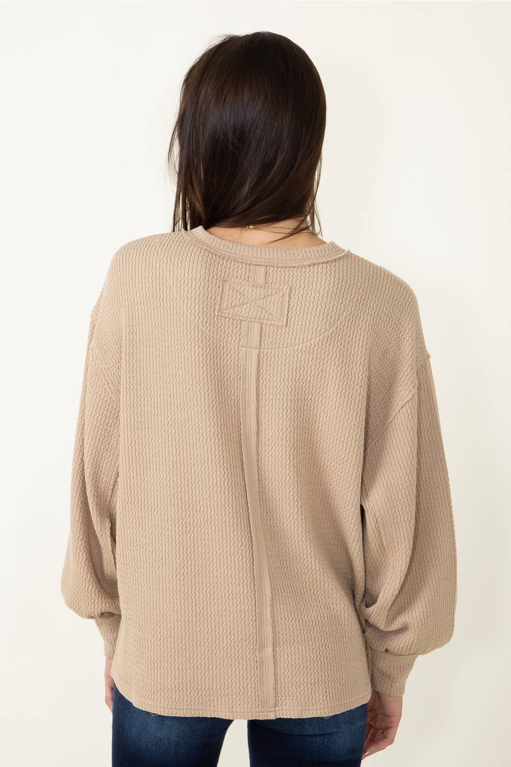 A woman viewed from the back wearing a beige waffle-knit sweater with a unique bow detail at the back neckline, paired with blue jeans against a plain white background.