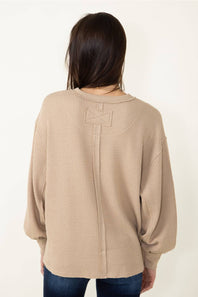 A woman viewed from the back wearing a beige waffle-knit sweater with a unique bow detail at the back neckline, paired with blue jeans against a plain white background.