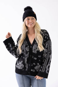 A woman smiling while wearing a black plush pom-pom beanie featuring a cuffed hem with a C.C. branded leather patch, paired with a black and white patterned cardigan.