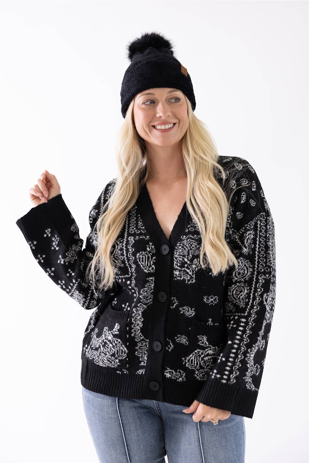 A woman smiling while wearing a black plush pom-pom beanie featuring a cuffed hem with a C.C. branded leather patch, paired with a black and white patterned cardigan.