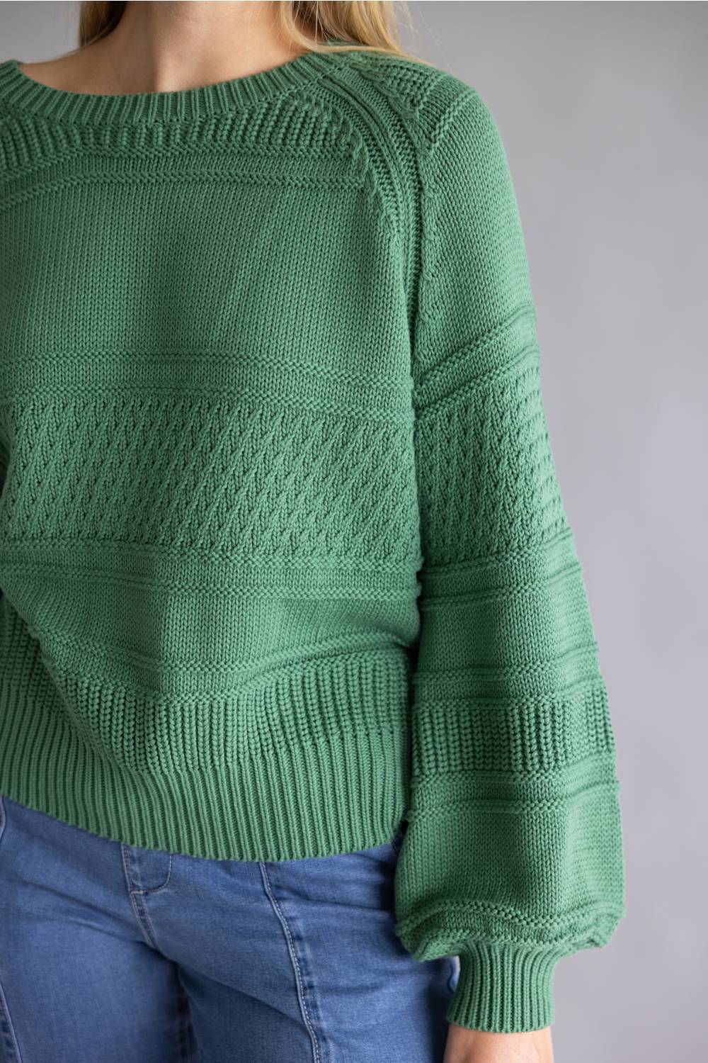 A person wearing a textured green knit sweater with blue jeans, shown from the shoulders to the hips against a plain gray studio background.
