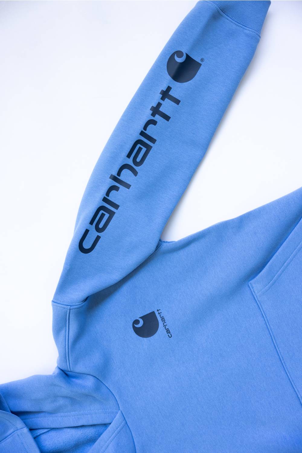 A close-up of a light blue Carhartt hoodie laid flat on a white background, showing the black Carhartt logo printed vertically on the sleeve and a smaller logo near the chest pocket.