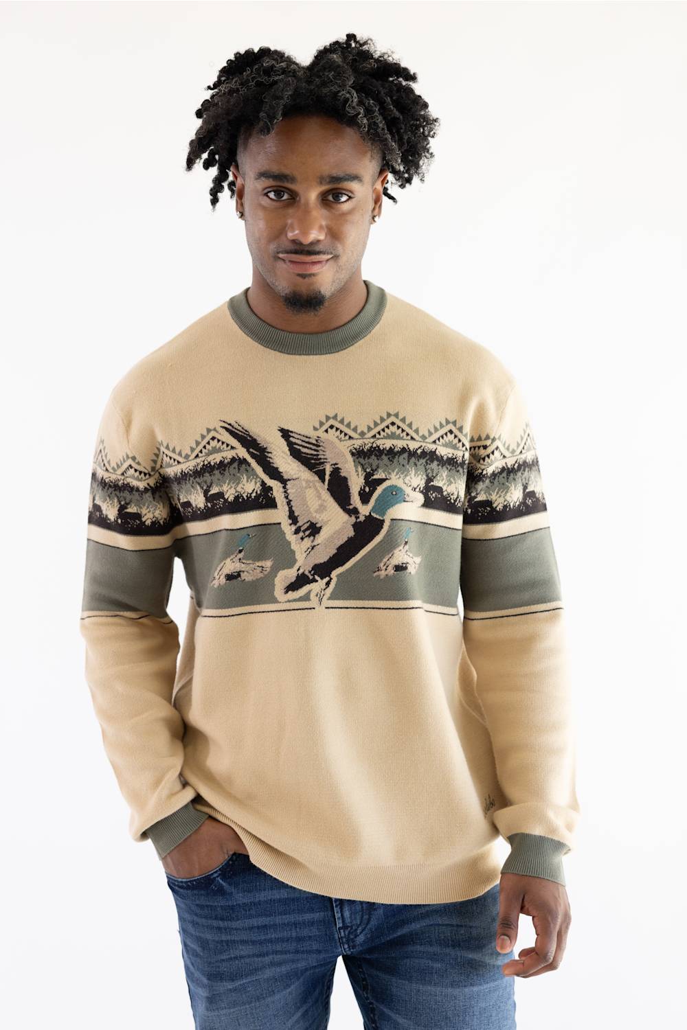 A man wearing a beige crewneck sweater with a graphic of a mallard duck and mountain patterns across the chest, paired with blue jeans, against a plain white background.