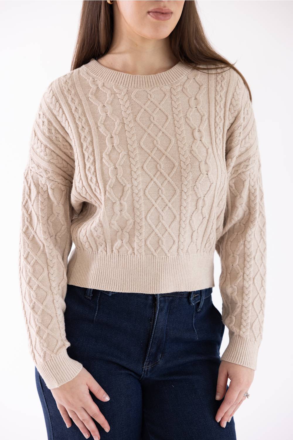 A woman wearing a beige cable-knit cropped sweater with dark blue jeans, shown from the nose down against a plain white background.