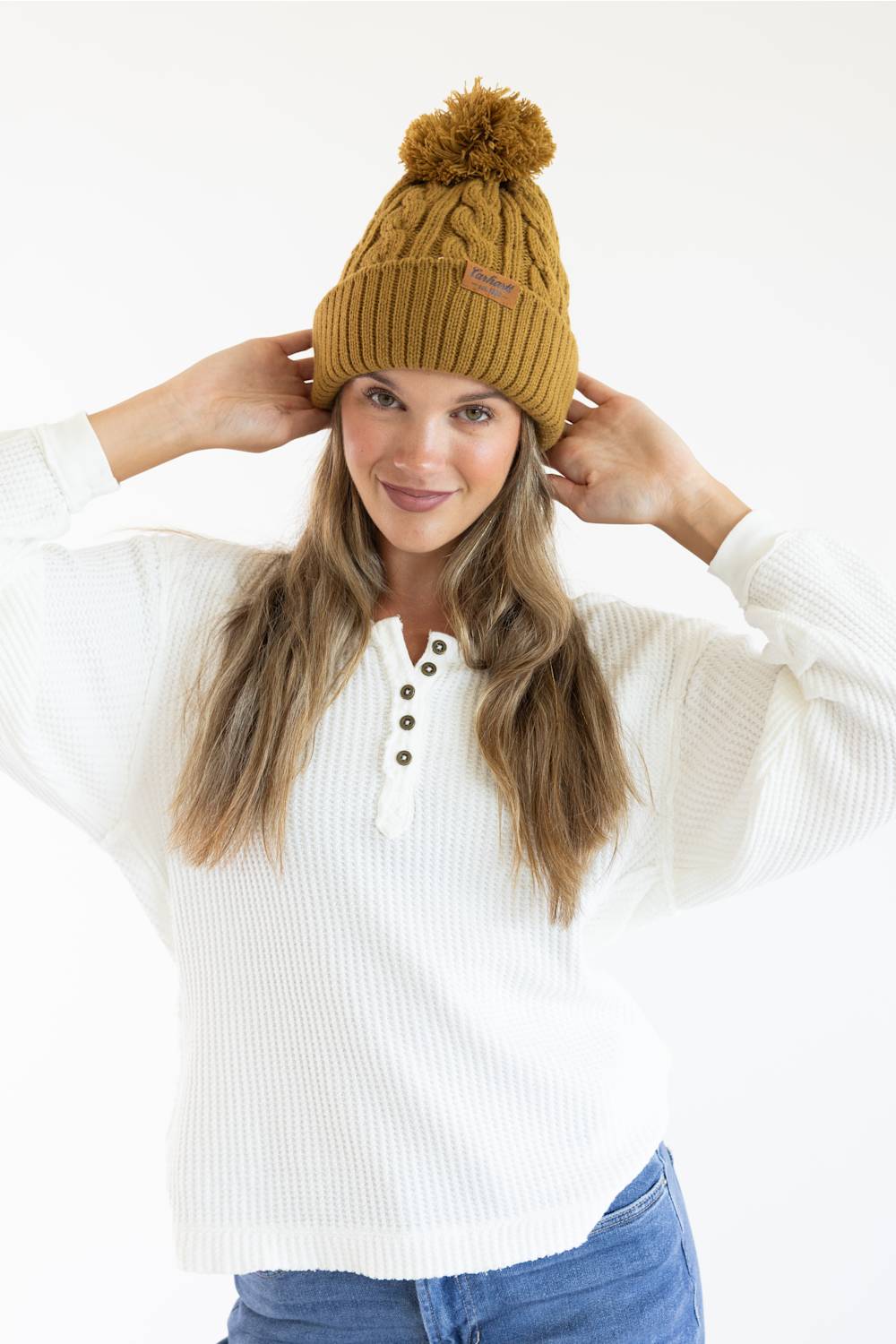 A woman wearing a mustard yellow cable-knit beanie with a pom-pom and a white ribbed henley sweater, posing against a white studio background.