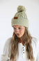 A woman wearing a light sage green cable-knit beanie with a matching pom-pom and a small brown leather brand patch. She is wearing a white henley knit sweater against a plain white studio background.