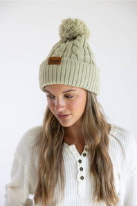 A woman wearing a light sage green cable-knit beanie with a matching pom-pom and a small brown leather brand patch. She is wearing a white henley knit sweater against a plain white studio background.