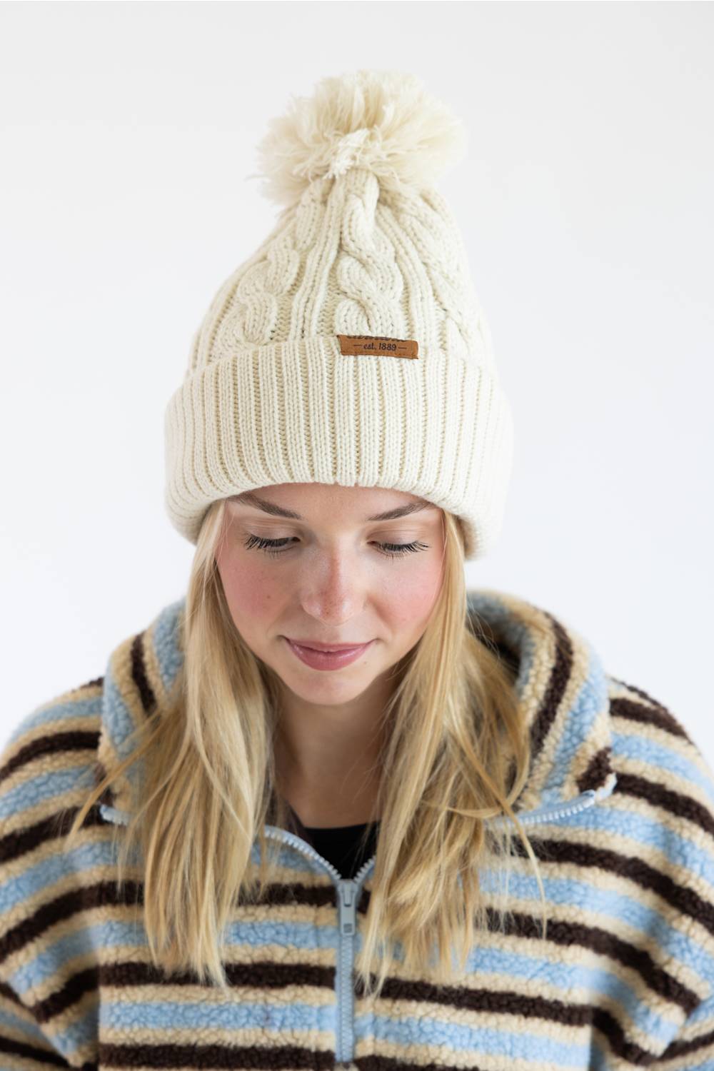 A woman wearing a cream cable-knit pom-pom beanie with a small brown leather brand tab, paired with a blue, brown, and cream striped fleece jacket, against a plain white background.
