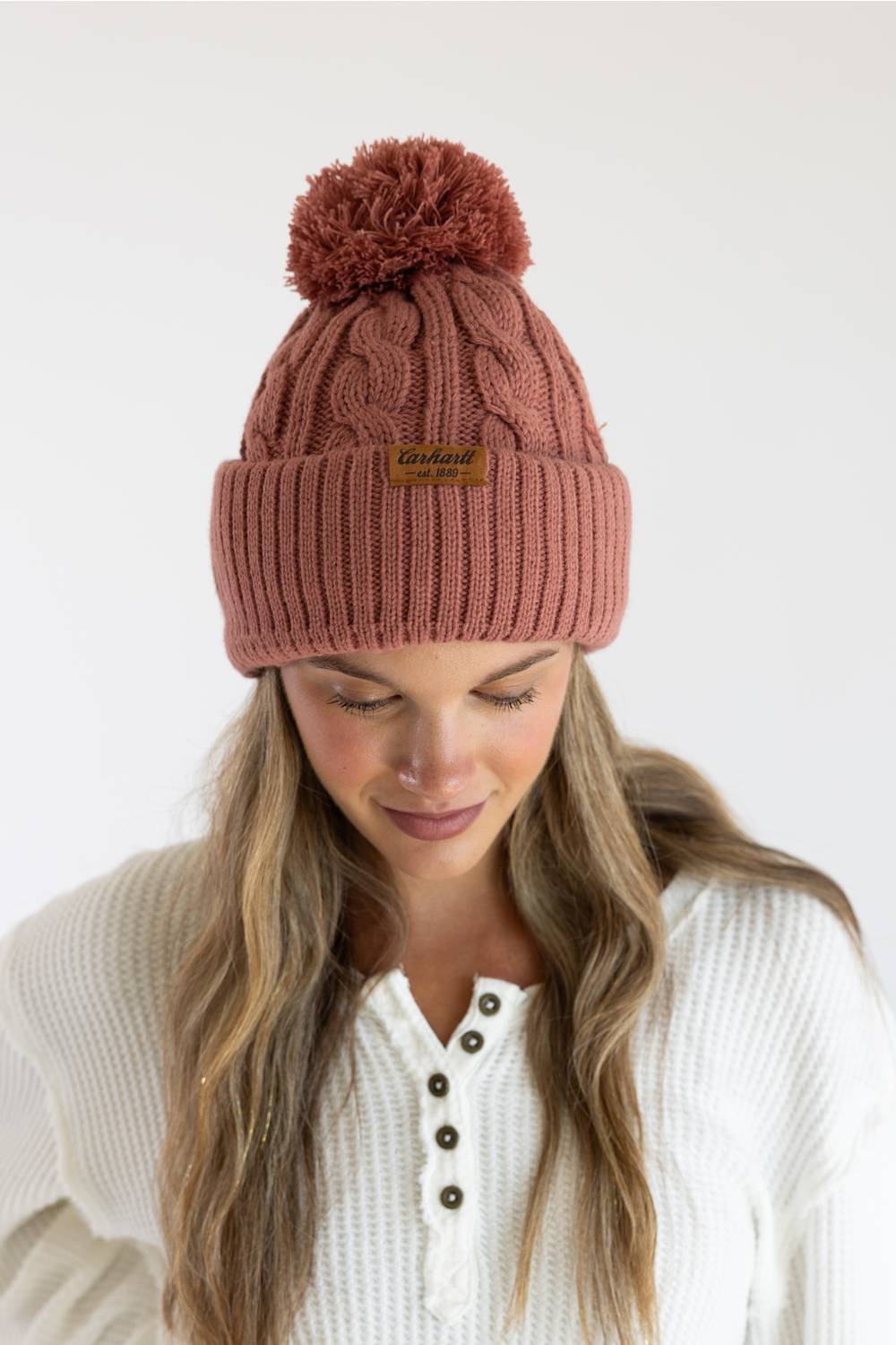 A woman wearing a dusty rose cable-knit beanie with a matching pom-pom and a small tan brand patch on the cuff. The background is a clean, neutral white studio backdrop.