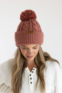 A woman wearing a dusty rose cable-knit beanie with a matching pom-pom and a small tan brand patch on the cuff. The background is a clean, neutral white studio backdrop.