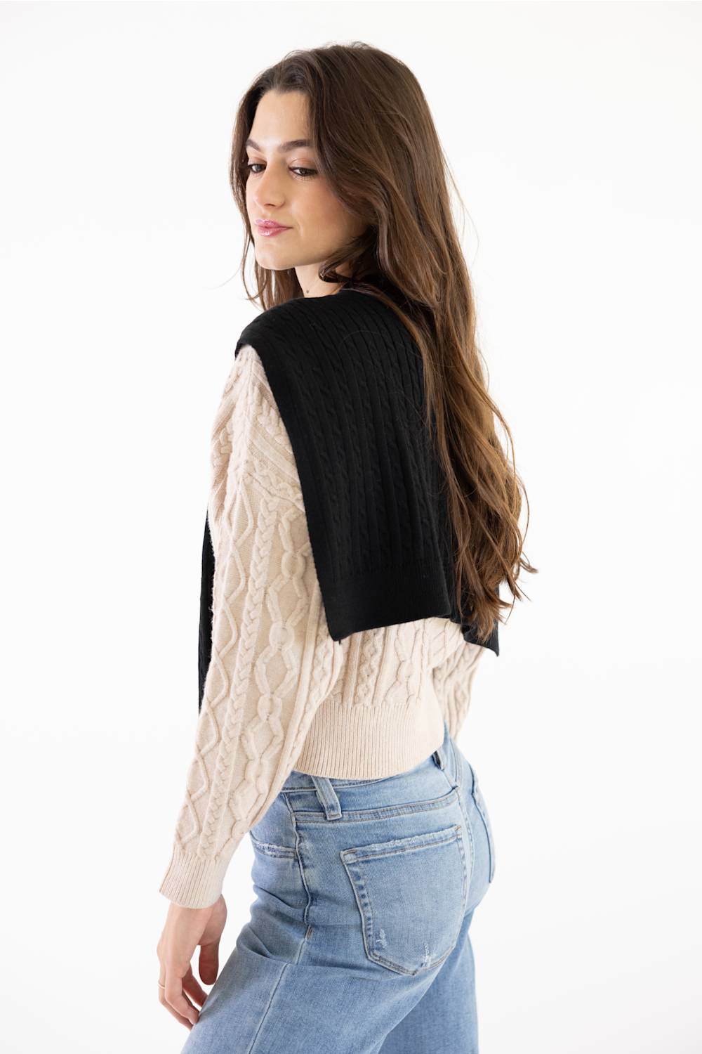 A woman with long brown hair is shown from the back wearing a black knit shrug or wrap over a cream cable-knit sweater and blue jeans, against a plain white studio background.