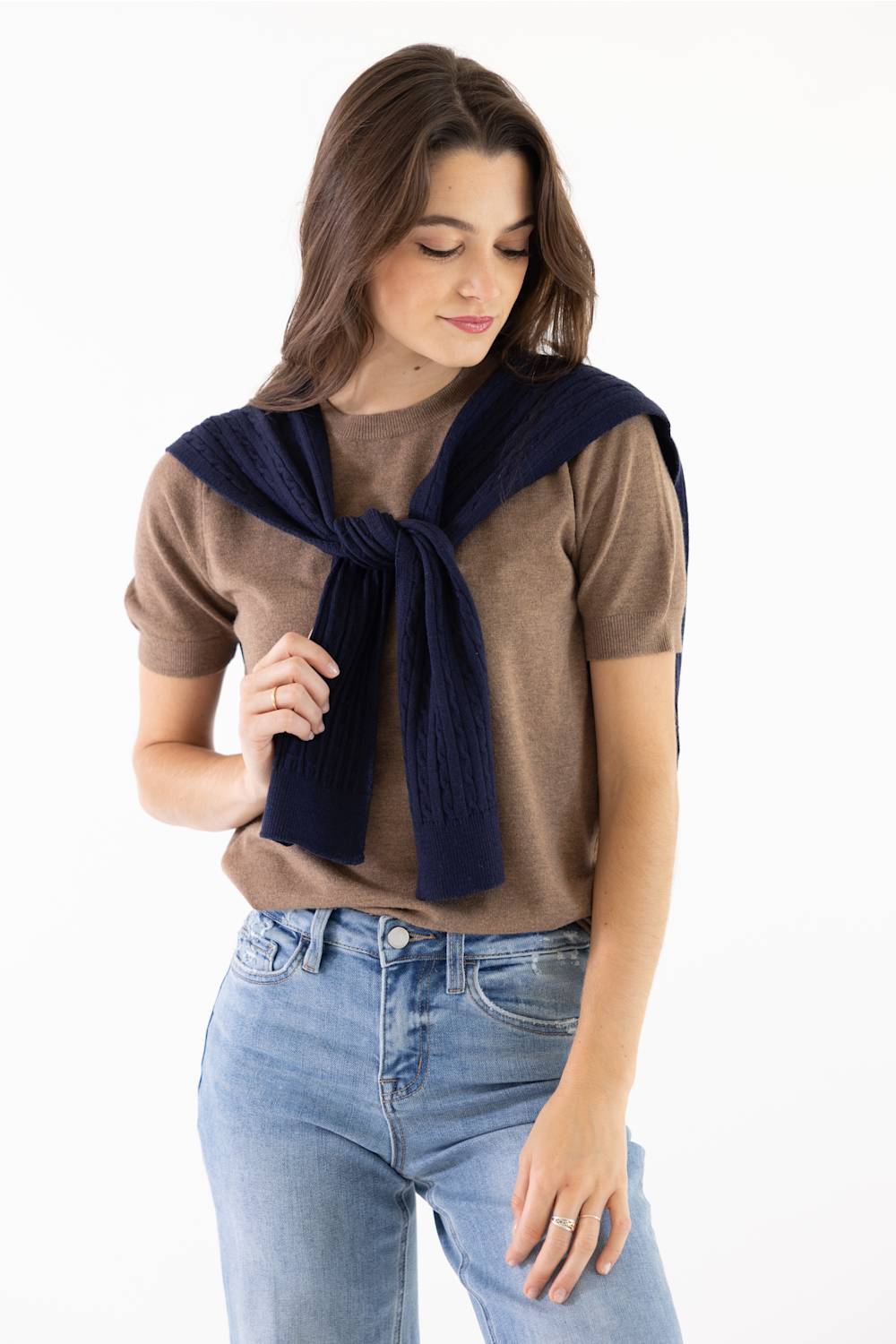 A woman wearing a brown short-sleeve t-shirt and blue jeans, with a navy blue knit sweater draped over her shoulders and tied in a knot at the front, against a plain white background.