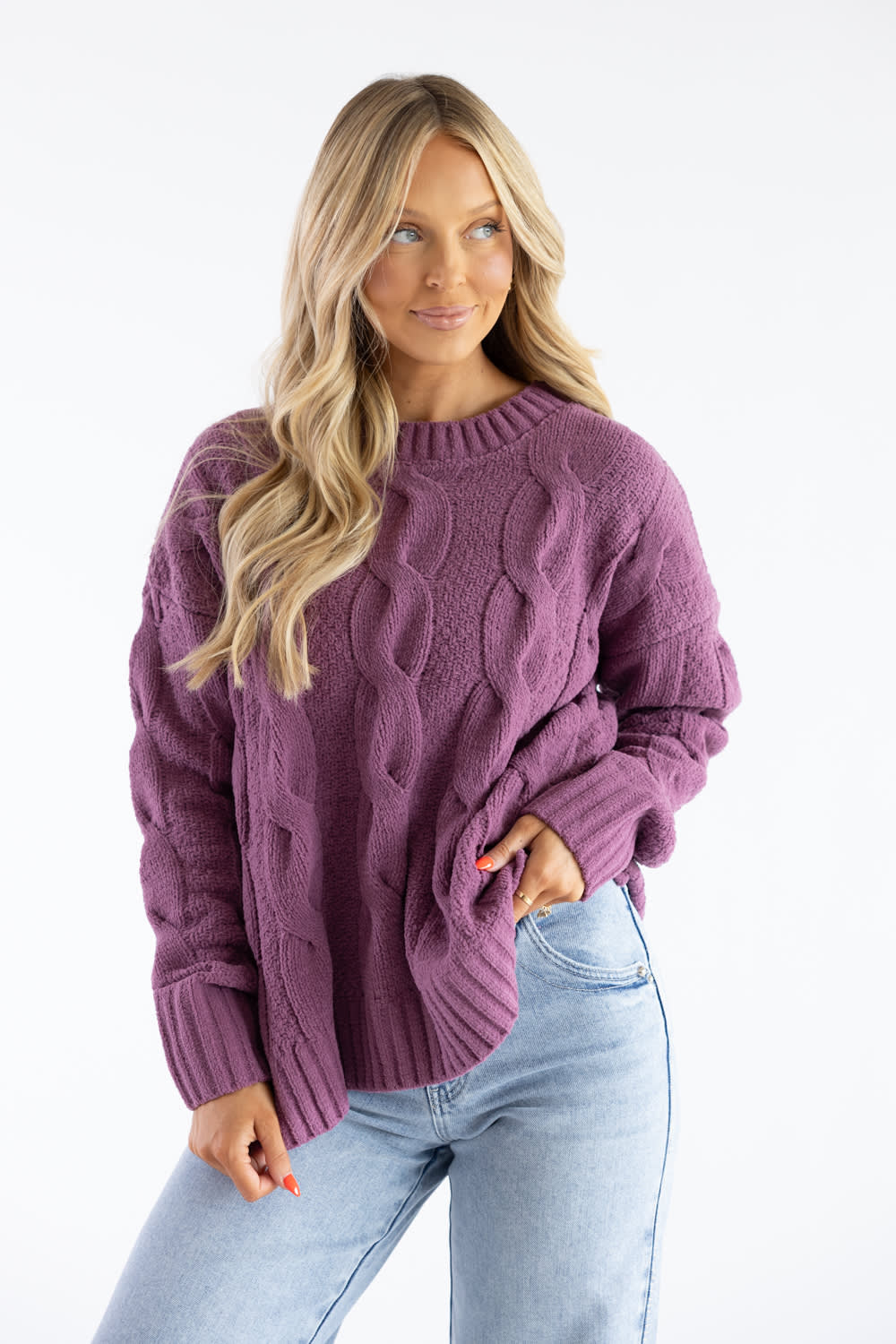 A woman wearing a chunky cable-knit sweater in a dusty purple color, paired with light-wash blue jeans, photographed against a clean white studio background.