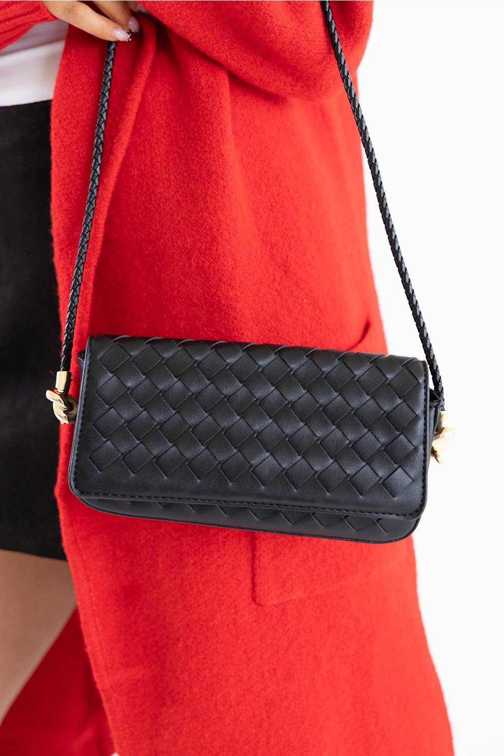 A black woven leather shoulder bag with a braided strap, shown worn over a bright red coat against a white background.