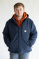 A man wearing a navy blue Carhartt hooded jacket over an orange hoodie and blue jeans, standing against a plain white background.