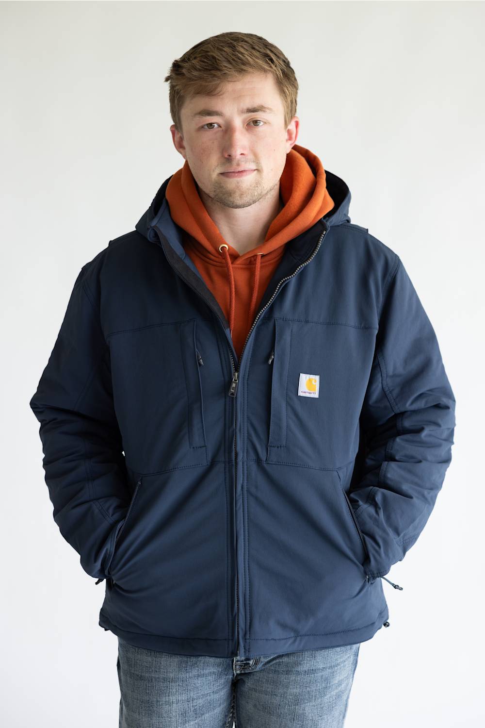 A man wearing a navy blue Carhartt hooded jacket over an orange hoodie and blue jeans, standing against a plain white background.